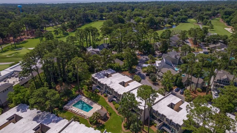 Aerial view of a golf-course residential community with white-roofed townhomes nestled among tall pine trees, a turquoise outdoor pool, winding streets and golf fairways stretching into the distance
