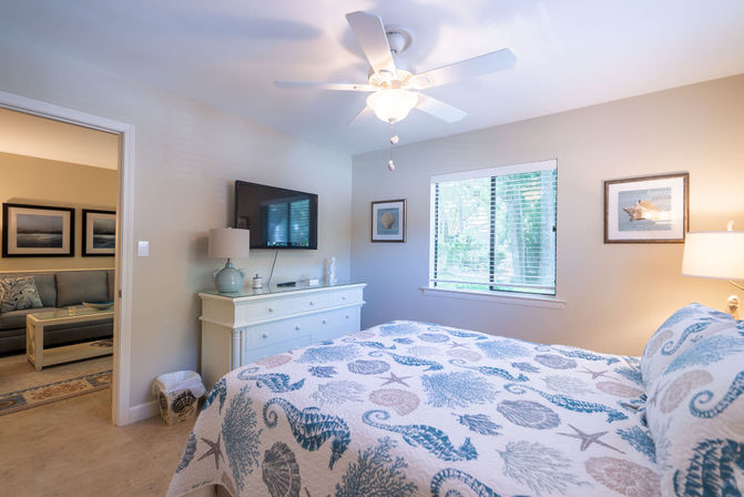 Coastal cottage bedroom with seahorse-and-shell quilt, white dresser and lamp, wall-mounted TV, ceiling fan, and window view of trees — bright beachy vibe.