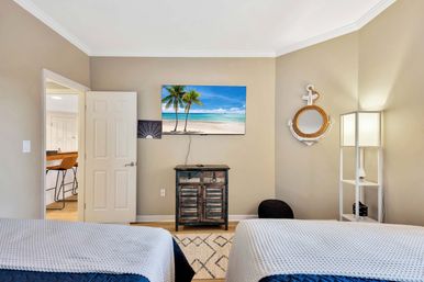 Coastal-themed bedroom with two twin beds, light beige walls, wall-mounted TV showing a tropical beach, rustic wooden cabinet, rope nautical wall decor, white floor lamp and open doorway to kitchen with bar stools.