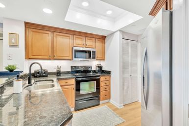 Bright, cozy kitchen with light wood cabinets, black granite countertops, undermount stainless sink, black electric range with microwave above, stainless steel refrigerator, recessed ceiling lights and light hardwood floor.