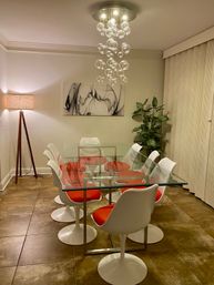Contemporary dining room with a glass-top table surrounded by white tulip chairs with red cushions, cascading bubble chandelier, tripod floor lamp, abstract black-and-white wall art, potted plant and tiled floor.