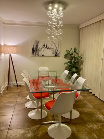 Contemporary dining room with a glass-top table surrounded by white tulip chairs with red cushions, cascading bubble chandelier, tripod floor lamp, abstract black-and-white wall art, potted plant and tiled floor.