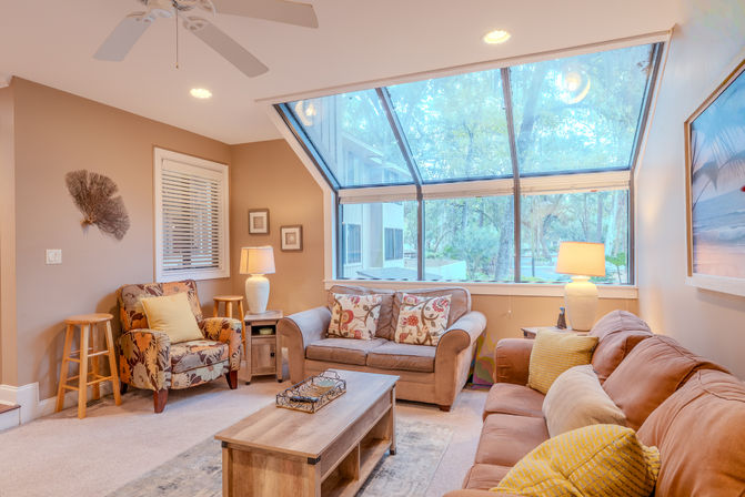 Cozy sunlit living room with a large angled skylight overlooking trees, tan sofas and patterned armchair, wooden coffee table, table lamps and yellow accent pillows.