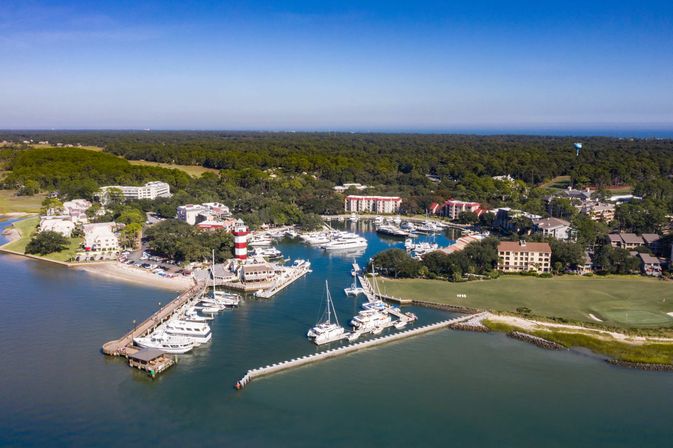 Aerial view of a sunny coastal marina with yachts and sailboats, a red-and-white striped lighthouse, docks, beachfront resorts, tree-lined shoreline and a nearby golf course.