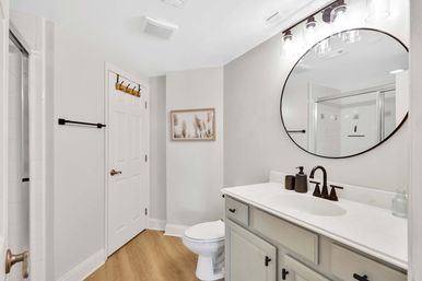 Bright, spa-like modern bathroom with a round mirror above a white single-sink vanity, bronze faucet, glass shower door, toilet, and wood-look flooring.