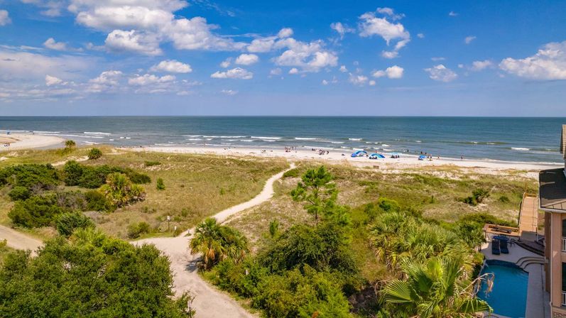 Aerial view of a sunny oceanfront beach with sandy dunes and a winding beach-access path, palm trees, small groups of beachgoers and umbrellas, gentle waves and a blue sky dotted with fluffy clouds.