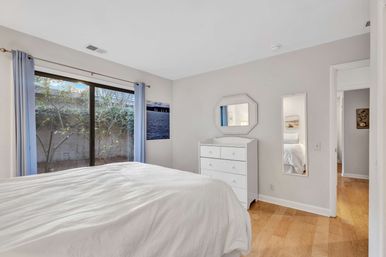 Bright cozy bedroom with white bedding, light hardwood floors, white dresser with octagonal mirror, full-length wall mirror, and a sliding glass door with blue curtains opening to a leafy garden view.