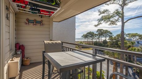 Sunny ocean-view balcony with a high-top table and chair, coastal pine trees and sandy shoreline visible beyond.