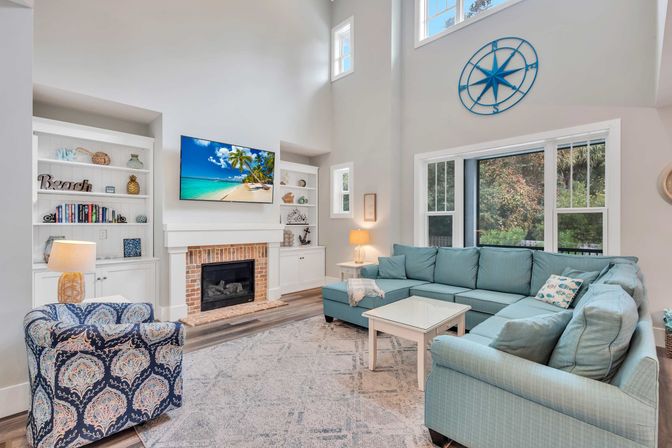 Bright beach-house living room with high ceilings, turquoise L-shaped sectional, patterned accent chair, brick fireplace with wall-mounted TV showing a tropical beach, built-in white shelves, large windows, nautical compass wall decor and light hardwood floors.