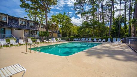 Sunny outdoor community swimming pool with inviting turquoise water, rows of white lounge chairs, townhouse facades and tall pine trees in the background.