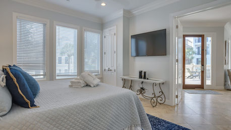 Bright coastal bedroom with gray quilted bed, navy throw pillows and folded towels, wall-mounted TV, decorative console table, and glass door opening to a sunny balcony with palm view.