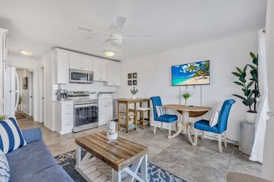 Bright coastal studio condo interior with open-plan white kitchen and stainless appliances, round wooden dining table with blue upholstered chairs, wall-mounted TV displaying a beach, blue sofa, wooden coffee table, tile floor, potted plant and ceiling fan.