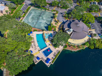 Aerial drone view of a lakeside community recreation complex with turquoise swimming pools, blue umbrellas and lounge chairs, adjacent tennis courts, and a round clubhouse with a conical roof surrounded by trees and parking