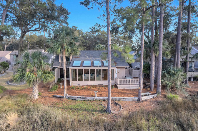 Coastal marshfront cottage with glass sunroom and three skylights, raised wooden deck among tall pine and palmetto palms, grassy tidal marsh in the foreground under a clear blue sky.
