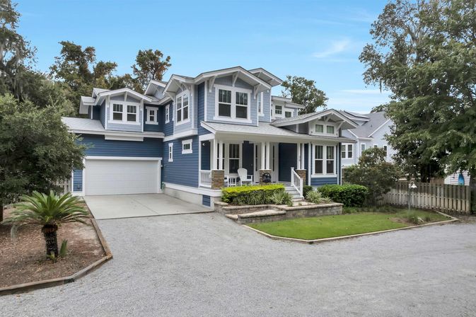 Two-story blue Craftsman-style house with white trim, covered front porch with chairs, attached two-car garage, raised stone planter, manicured lawn, gravel driveway and mature trees