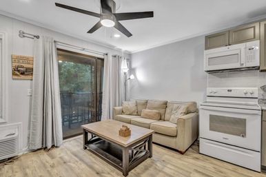 Cozy open-plan apartment living area with beige sofa, wooden coffee table, white stove and microwave, ceiling fan, floor lamp, and sliding glass door opening to a tree-lined balcony; light wood-look flooring and neutral decor.