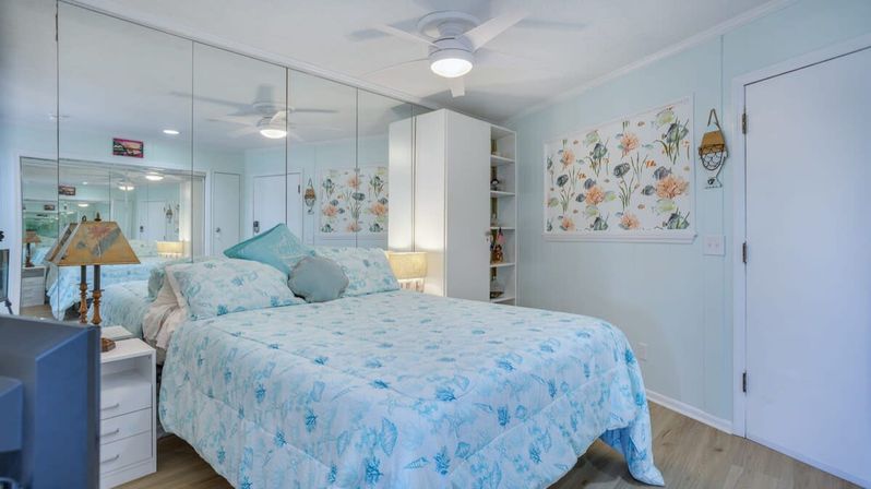 Cozy beachy bedroom with light-blue shell-pattern bedding on a neatly made bed, mirrored wall reflecting the room, ceiling fan, floral wall art, white bedside tables and shelving, and light wood floors.
