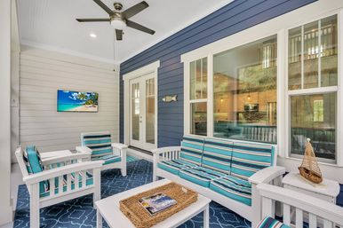Bright coastal-style screened porch with white wooden seating, turquoise-striped cushions, navy patterned rug, wall-mounted TV showing a beach, ceiling fan, wicker tray and decorative sailboat beside large windows