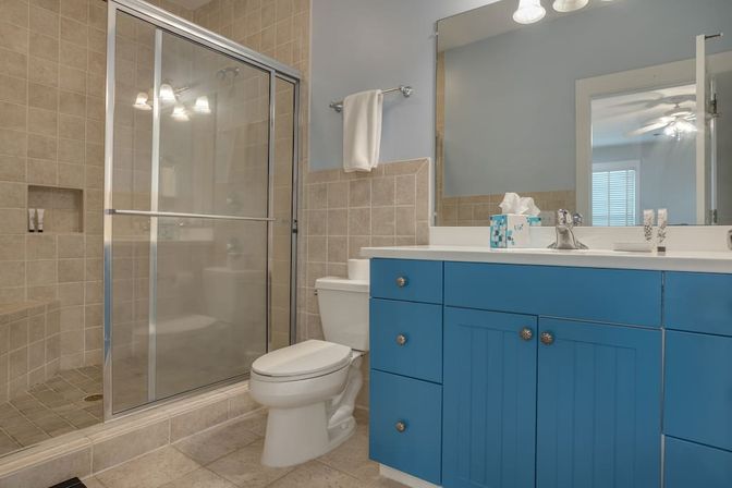 Cheerful modern bathroom with blue shaker vanity and white countertop, tiled walk-in shower with glass sliding doors, white toilet, large mirror and towel on rack