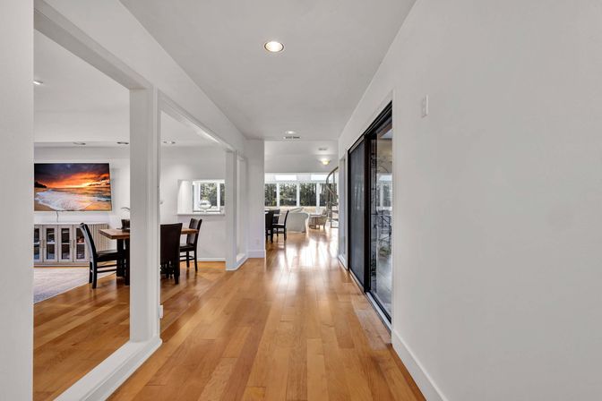 Inviting sunlit open-plan hallway with warm hardwood floors, sliding glass doors, dining table, wall-mounted TV showing a sunset beach scene and a spiral staircase in the background.