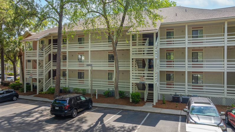 Sunny, tree-lined three-story apartment building with exterior wooden walkways and staircases, parked cars in the lot — suburban condo exterior scene.