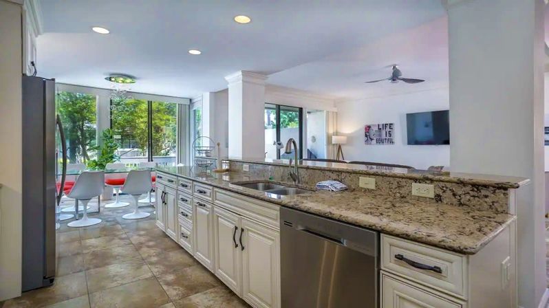 Sunlit modern open-concept kitchen with granite countertops, white cabinets, stainless steel dishwasher and breakfast bar overlooking a dining nook with red chairs and large windows showing leafy outdoor views.