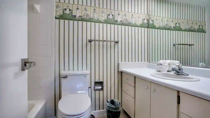 White coastal-style bathroom with vertical striped wallpaper and lighthouse border, white toilet beside a tub, long vanity with sink and mirror, towel racks and small trash bin.