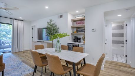 Airy modern dining area in a coastal-style apartment: white marble table with six tan leather chairs, glass vase of green leaves, built-in coffee bar and shelves, wood floors, sliding glass door to a leafy balcony, and a bathroom with striped tile visible.