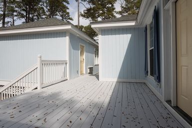 Sunlit light-blue cottage exterior with white railing and wide gray wooden deck strewn with leaves