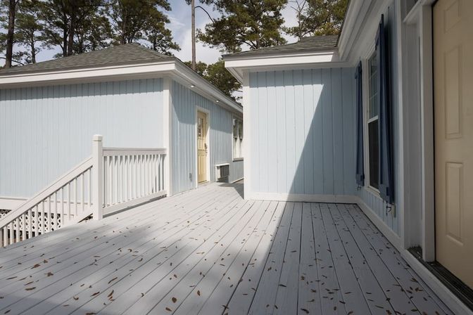 Sunlit light-blue cottage exterior with white railing and wide gray wooden deck strewn with leaves