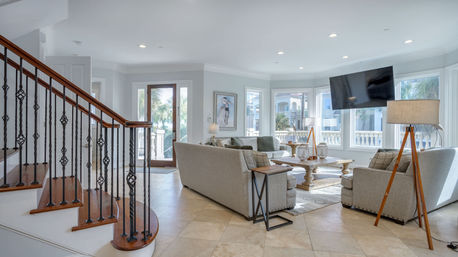 Bright coastal living room with neutral sofas, wooden coffee table, tripod floor lamp, wall-mounted TV and wrought-iron staircase, large windows revealing palm trees and balcony