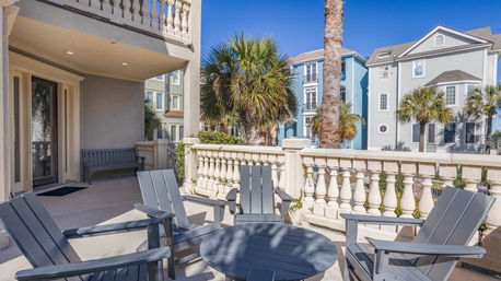 Sunny seaside condo patio with gray Adirondack chairs around a round table, ornamental stone balustrade, palm trees and pastel beach houses under a bright blue sky.