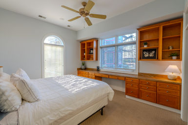 Cozy sunlit bedroom with white duvet, arched window, ceiling fan, and long built-in wooden desk and shelving with drawers and lamp.