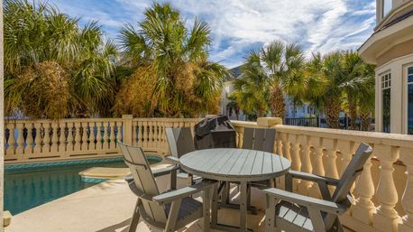 Sunlit tropical poolside patio with round gray table, four matching chairs and a covered grill beside a small pool, stone balustrade and palm trees under a blue sky.