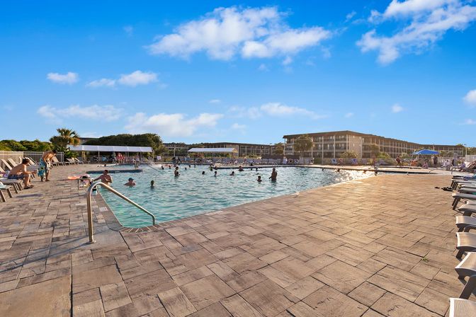 Sunny coastal resort pool with families swimming, palm trees and lounge chairs on a spacious tiled deck under a bright blue sky