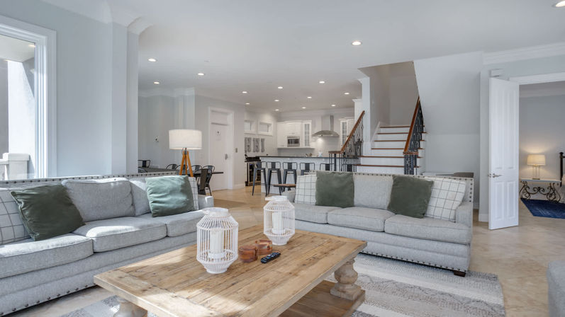 Bright, open-concept living room in a modern home with two gray sofas and green throw pillows, rustic wooden coffee table with decorative white lanterns, adjoining kitchen island with bar stools, and a staircase leading upstairs.