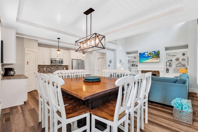 Coastal open-concept beach house interior with a large square wooden dining table surrounded by white chairs, geometric pendant light, adjoining white kitchen with stainless steel appliances and mosaic backsplash, blue sofa, built-in shelving, wall TV showing a beach scene, and wood-look floors.