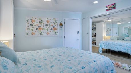 Cozy beach-style bedroom with light-blue seashell quilt on the bed, tropical fish and coral wall art, mirrored sliding closet doors, and light wood flooring.