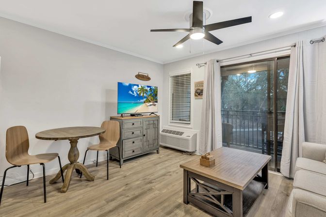 Cozy coastal-style vacation rental living room with wood-look floors, round bistro table and two chairs, dresser with TV showing a tropical beach, coffee table, ceiling fan and sliding glass door to a tree-lined balcony.