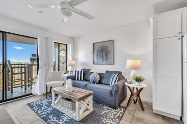 Sunlit beachfront condo living room with navy slipcovered sofa, striped pillows, rustic wood coffee table, coral-pattern rug and sliding glass doors to an ocean-view balcony.