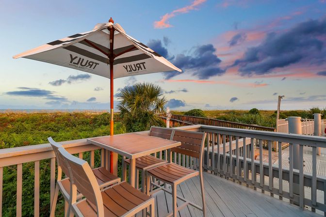 Beachfront deck at sunset with high-top table, chairs and umbrella overlooking coastal dunes, palm plants and ocean horizon