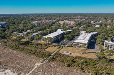 Aerial view of beachfront condominiums nestled behind coastal dunes and marshland with palm trees, parking areas and a wooded residential neighborhood stretching to the horizon under a clear blue sky.