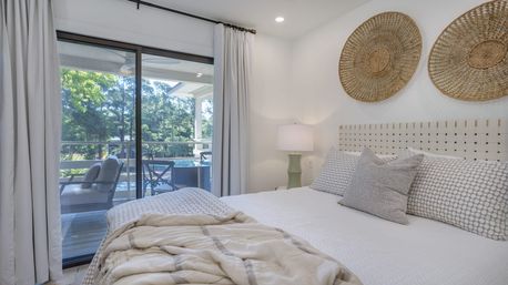 Bright coastal-style bedroom with sliding glass doors to a patio and pool, neutral linens, woven wall baskets and outdoor seating visible through glass.