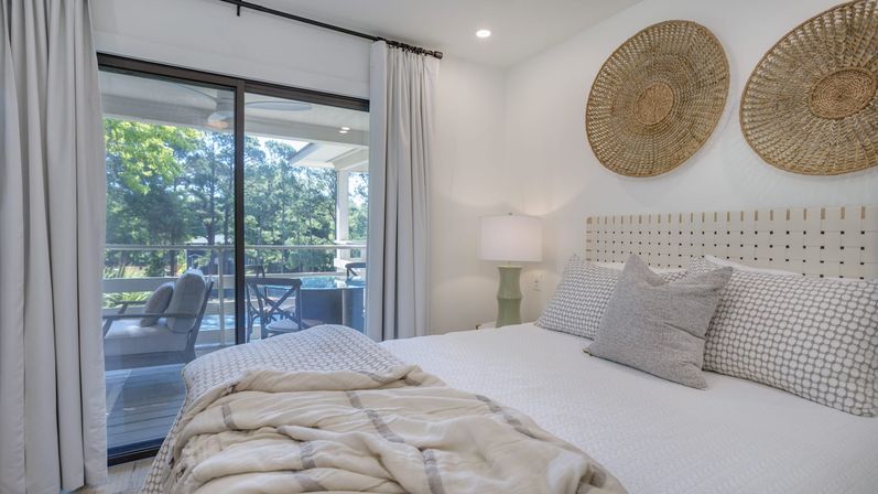 Bright coastal-style bedroom with sliding glass doors to a patio and pool, neutral linens, woven wall baskets and outdoor seating visible through glass.
