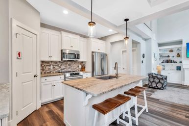 Bright open-concept modern kitchen with white cabinets, marble-topped island and wooden bar stools, stainless steel appliances, tile backsplash, pendant lights and hardwood floors.