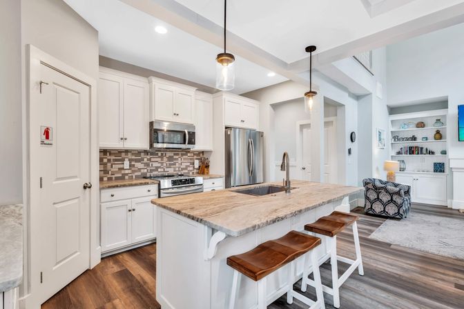 Bright open-concept modern kitchen with white cabinets, marble-topped island and wooden bar stools, stainless steel appliances, tile backsplash, pendant lights and hardwood floors.