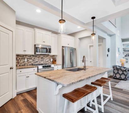 Bright open-concept modern kitchen with white cabinets, marble-topped island and wooden bar stools, stainless steel appliances, tile backsplash, pendant lights and hardwood floors.