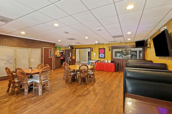 Bright, cozy restaurant interior with hardwood floors, round wooden tables and wicker chairs, black leather booths, recessed ceiling lights, wall TVs and a red-skirted buffet table.