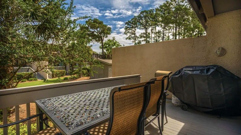 Sunny residential balcony patio with metal lattice dining table, woven chairs and covered grill overlooking pine trees and a shared lawn under a blue sky.