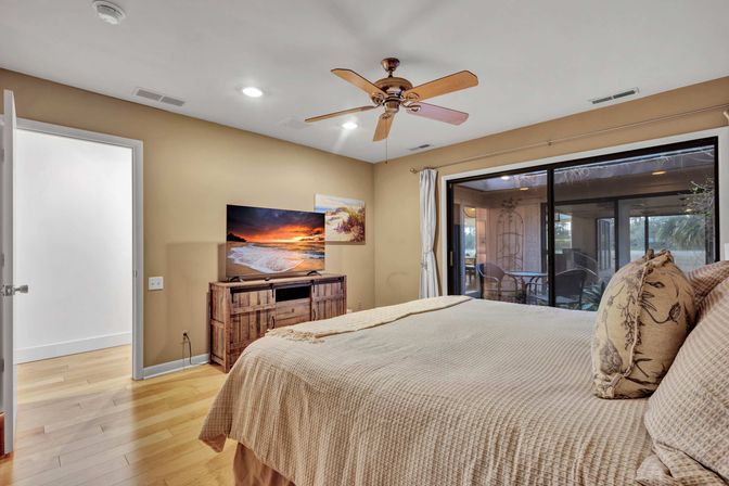 Cozy master bedroom with light wood floors, beige textured bedding and patterned pillows, ceiling fan, flat-screen TV on a rustic wooden console, and large sliding glass door to a screened patio.